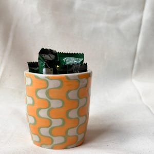 May include: A ceramic cup featuring a repeating pattern of orange and light green interlocking wave shapes. The cup is filled with several small, dark green rectangular packets with serrated edges, partially visible at the top opening.