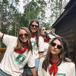 Pu&ograve; includere: Quattro persone con occhiali da sole, bandane rosse e magliette color crema con la scritta "Camp Raines" e una grafica di un albero verde. Sono in piedi su un ponte di legno con alberi sullo sfondo.