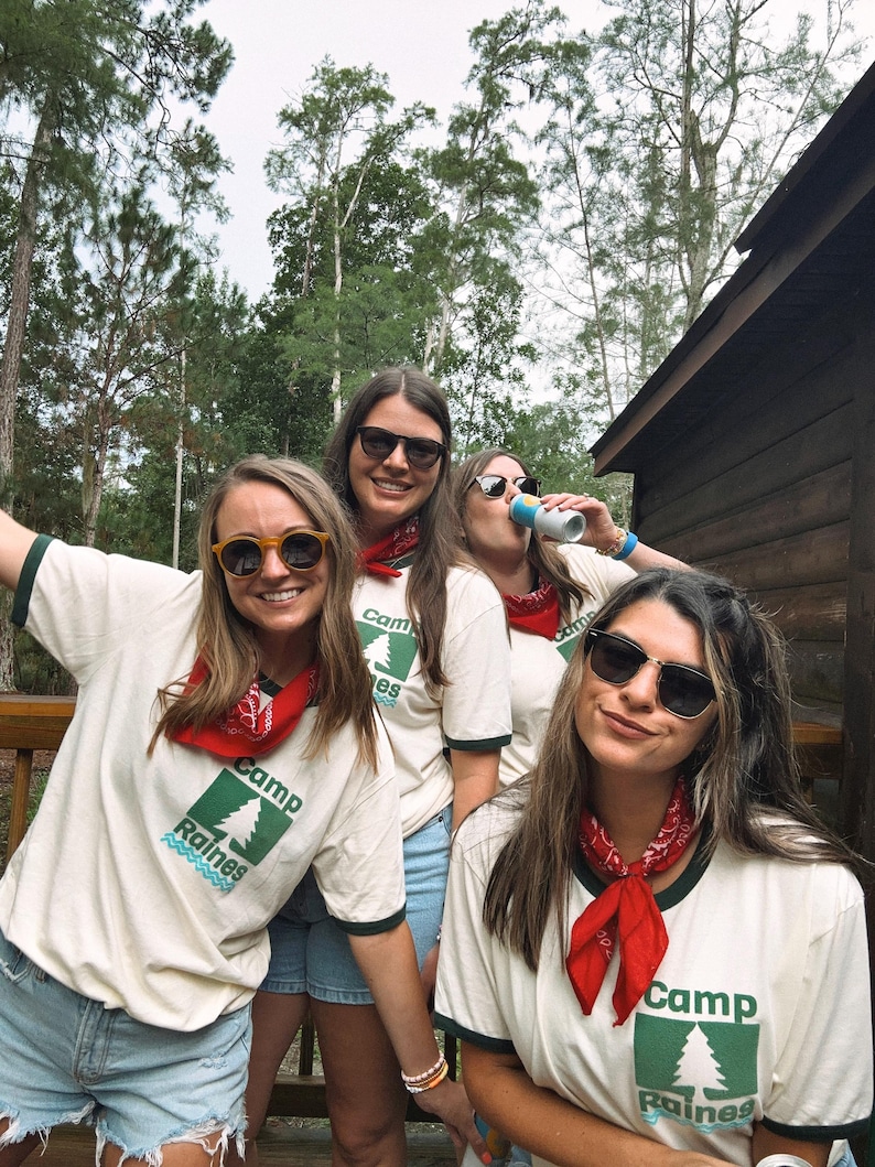 Pu&ograve; includere: Quattro persone con occhiali da sole, bandane rosse e magliette color crema con la scritta "Camp Raines" e una grafica di un albero verde. Sono in piedi su un ponte di legno con alberi sullo sfondo.
