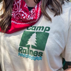 Pu&ograve; includere: T-shirt color crema con testo grafico verde e blu "Camp Raines". La maglietta ha un bordo verde sul colletto e sulle maniche. Una bandana rossa con un motivo paisley bianco &egrave; indossata intorno al collo.