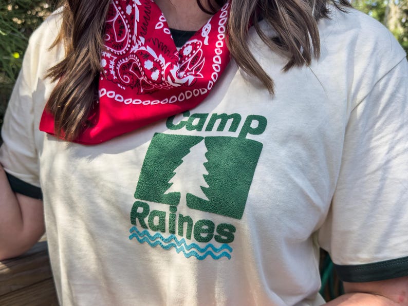 Pu&ograve; includere: T-shirt color crema con testo grafico verde e blu "Camp Raines". La maglietta ha un bordo verde sul colletto e sulle maniche. Una bandana rossa con un motivo paisley bianco &egrave; indossata intorno al collo.