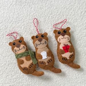 May include: Three felt otter ornaments with red and white string loops. One otter wears a green scarf, one holds a white shell, and one holds a small red gift box. Each brown and tan otter has black bead eyes and stitched facial details.