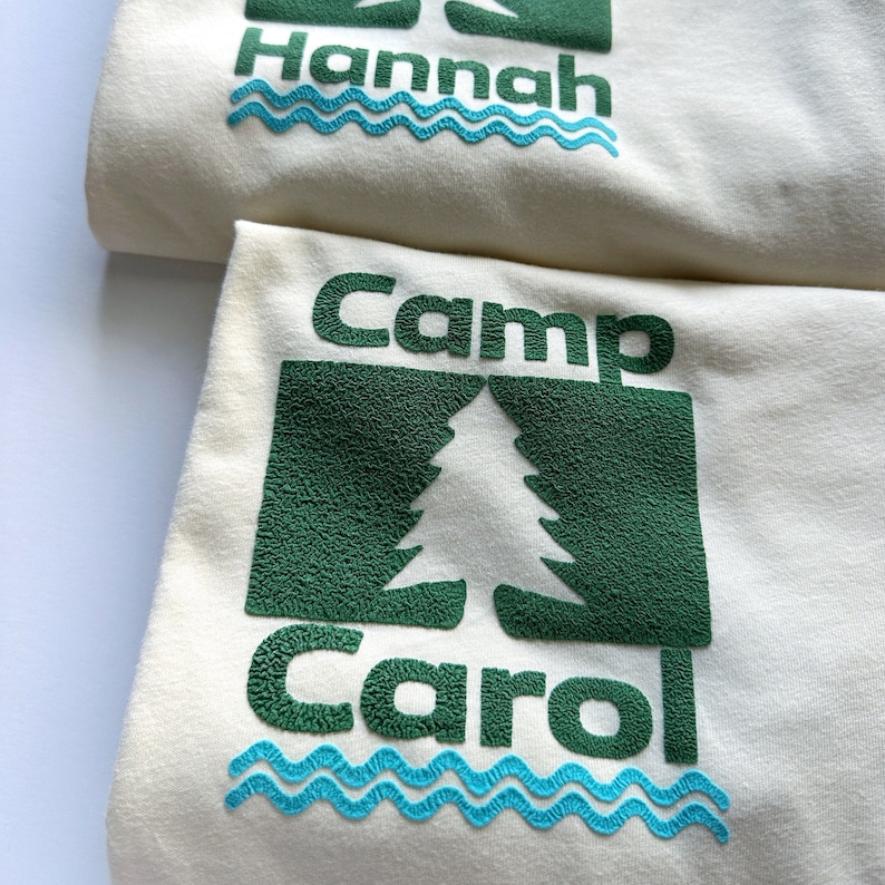Pu&ograve; includere: Due felpe color crema con disegni verdi e blu. Una dice "Hannah" sopra una linea ondulata blu. L'altra dice "Camp Carol" con una grafica ad albero verde e una linea ondulata blu. Il testo e la grafica hanno un aspetto strutturato.