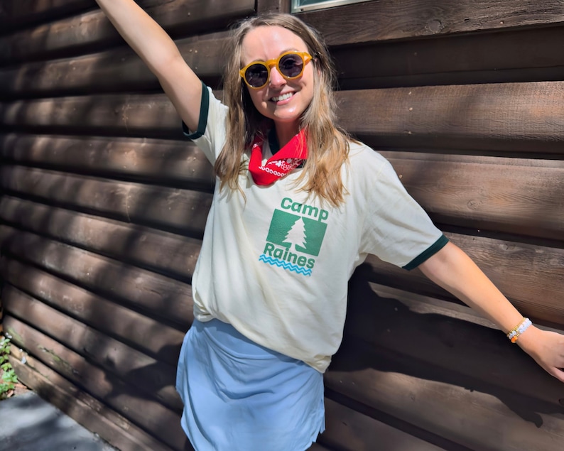 May include: A person wearing a white t-shirt with a green and blue logo that says 'Camp Raines' and a red bandana around their neck. They are wearing yellow sunglasses and a blue skirt.