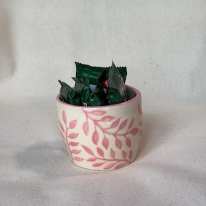May include: A small, cream-colored ceramic cup featuring a hand-painted pink leaf pattern around the exterior and a pink rim. The cup is filled with several small, dark green, individually wrapped rectangular food items.