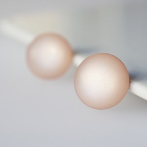 May include: A pair of round, blush-colored stud earrings. The earrings have a matte finish and are set against a white surface. The soft pink hue of the earrings provides a subtle, elegant look.