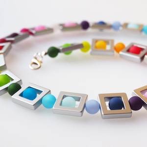 May include: A rainbow colored necklace with silver square beads and round glass beads. The necklace is made of a variety of colors, including red, orange, yellow, green, blue, indigo, and violet.