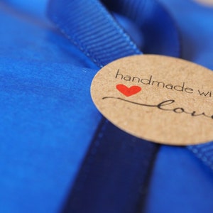 Puede incluir: Una etiqueta redonda de papel marr&oacute;n con el texto "handmade with love" en letras negras y un coraz&oacute;n rojo. La etiqueta est&aacute; atada a una cinta azul.
