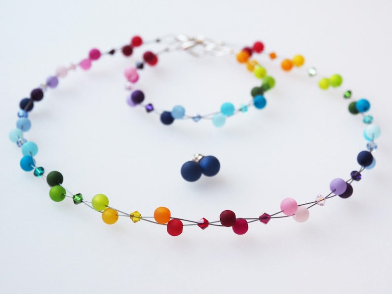 May include: A rainbow colored beaded necklace with matching earrings. The necklace is made of small, round beads strung on a thin wire. The beads are in a rainbow order, starting with red and ending with purple. The earrings are also made of small, round beads in the same rainbow order.