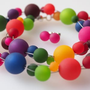 May include: A colorful necklace made of large, round, translucent beads in various shades of red, orange, yellow, green, blue, purple, and brown. The beads are strung on a silver wire and are arranged in a circular pattern. The necklace is accompanied by a pair of matching earrings.