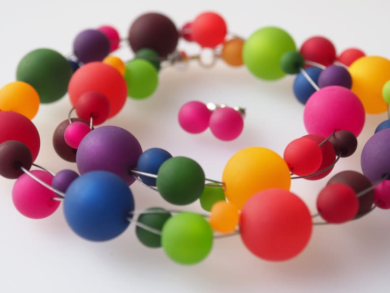 May include: A colorful necklace made of large, round, translucent beads in various shades of red, orange, yellow, green, blue, purple, and brown. The beads are strung on a silver wire and are arranged in a circular pattern. The necklace is accompanied by a pair of matching earrings.