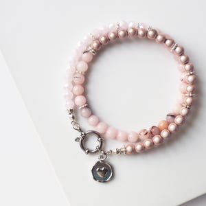 May include: A multi-strand beaded bracelet featuring a mix of pale pink, rose gold, and translucent faceted beads. The bracelet is secured with a silver-toned circular clasp and includes a small, round silver-toned charm with a heart cutout.