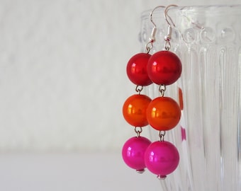May include: A pair of dangle earrings with three round beads in shades of red, orange, and pink. The beads are glossy and have a metallic finish.