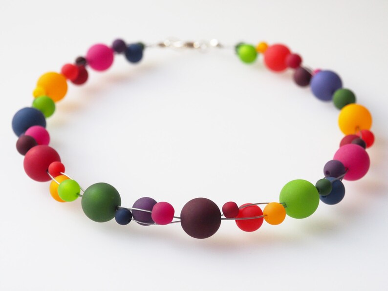 May include: A colorful necklace made of round beads in various shades of red, orange, yellow, green, blue, and purple. The beads are strung on a silver wire.