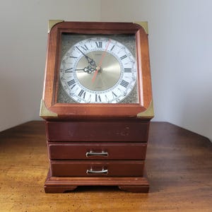 May include: A wooden clock with a gold-colored face and Roman numerals. The clock is encased in a rectangular wooden frame with brass corner accents. Below the clock face are three small drawers with silver-colored handles. The clock sits on a wooden base.