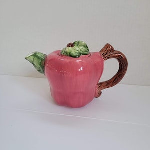 Puede incluir: Una tetera de cerámica rosa con forma de manzana con una hoja verde y un asa marrón.