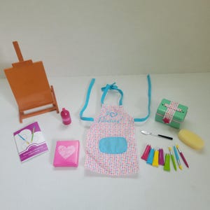 Könnte beinhalten: Eine Sammlung von Miniatur-Kunstzubehör, darunter eine orangefarbene Staffelei, eine rosa Farbflasche und eine gepunktete Schürze mit dem Text "I'm Painting!". Ebenfalls abgebildet sind Farbtuben, Pinsel, ein Palettenmesser und eine kleine Geschenkbox.