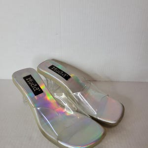 May include: A pair of clear, iridescent Mudd brand sandals with a square toe and clear heels. The sandals have a silver footbed and the Mudd logo is visible on the insole.