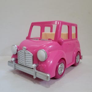 May include: A bright pink toy car with a classic design. The car features silver-colored accents on the front bumper, headlights, and grill. The wheels are pink with silver hubcaps. The car has a light tan interior.