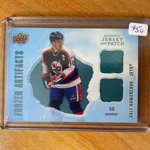Könnte beinhalten: Eine Hockey-Sammelkarte mit Dale Hawerchuk von den Jets. Die Karte enthält einen Trikotaufnäher und den Text "Frozen Artifacts". Die Karte ist in einem Schutzhalter eingeschlossen.