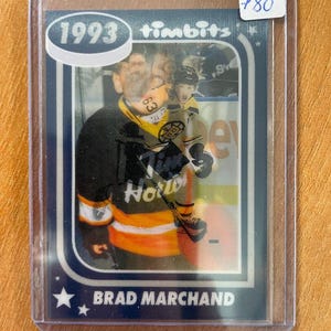 Puede incluir: Una tarjeta de hockey Timbitz de 1993 con Brad Marchand con el uniforme de los Boston Bruins. La tarjeta está dentro de una funda protectora, con un diseño azul y blanco y el año 1993 en la parte superior.