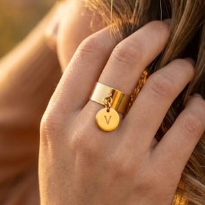 Peut inclure: Une bague anneau en métal doré avec une surface large et lisse, accompagnée d'une petite breloque circulaire suspendue. La breloque est gravée de la lettre majuscule V, qui est clairement visible au centre du disque. La bague est polie.