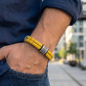 Può includere: Un bracciale in pelle intrecciata gialla a doppio filo con una chiusura cilindrica in metallo caratterizzata da un motivo a punti testurizzato. Il bracciale è indossato al polso, mettendo in risalto il colore giallo vivace dei cordini in pelle.