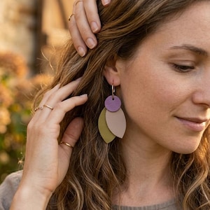 May include: Earrings featuring a purple disc and two leaf-shaped pendants in olive green and beige, suspended from gold-coloured hoops. The image shows a person wearing the earrings.