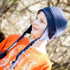May include: A navy blue fleece hat with braided ties, worn by a person. The hat has ear flaps and a rounded crown. The person is wearing an orange long-sleeved shirt with a graphic design. The background is blurred with natural light.