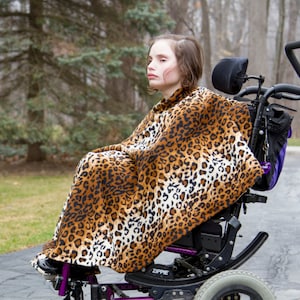 May include: A young person with brown hair and a leopard print blanket is sitting in a wheelchair. The wheelchair has a purple headrest and a black frame.