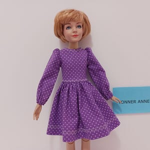May include: A doll wearing a purple dress with white polka dots. The dress has long sleeves and a flared skirt. The doll has short, light brown hair and is holding a blue sign that reads "YONNER ANNE".
