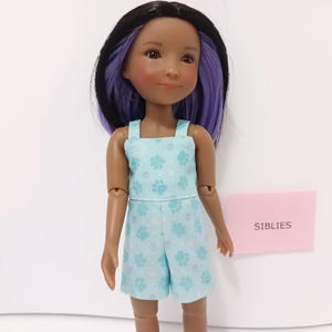 May include: A doll with dark skin and straight hair that is black on top and purple underneath. The doll wears a light blue romper featuring a pattern of small white dots and teal paw prints. A small pink rectangular sign reads SIBLIES.