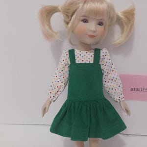 May include: A doll with blonde hair styled in two pigtails, wearing a long-sleeved white shirt featuring a multicolored polka dot pattern under a solid dark green corduroy jumper dress. A small pink rectangular sign displays the text SIBLIES.