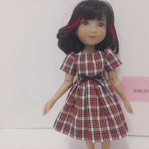 May include: A doll with dark hair featuring red highlights, wearing a short-sleeved dress with a red, white, and black plaid pattern and a thin black ribbon at the waist. A small pink rectangular sign with the text SIBLIES is visible.