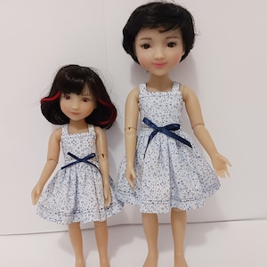 May include: Two dolls wearing matching blue and white floral print dresses with navy blue sashes. The dolls have black hair and are standing on a white background.
