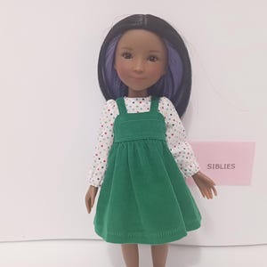 May include: A doll with dark hair featuring purple highlights, wearing a long-sleeved white shirt with a multicolored polka dot pattern under a solid green corduroy pinafore dress. A small pink sign with the text SIBLIES is visible.