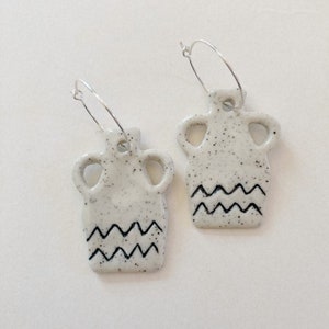 May include: A pair of white ceramic earrings shaped like a vase with a black wavy line design. The earrings are attached to silver hoop earrings.