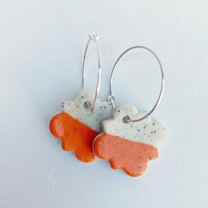 Könnte beinhalten: Ein Paar silberne Creolen mit orangefarbenen und weißen Keramik-Blumenanhängern. Die Blumen haben eine gesprenkelte weiße Glasur und eine glatte orangefarbene Glasur.