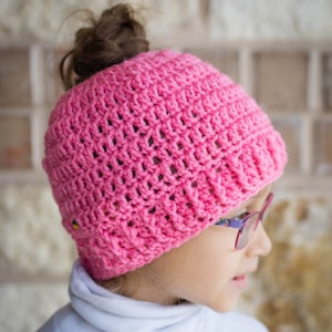 May include: A pink crocheted hat with a hole in the top for a ponytail. The hat is made with a simple stitch pattern and has a soft, textured look.