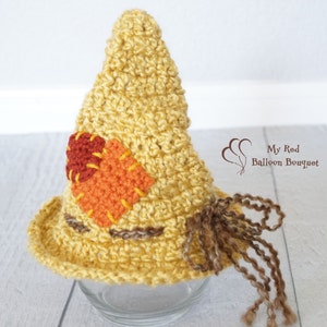 May include: A yellow crocheted hat with a pointed top, featuring a square orange patch and a circular dark red patch stitched on the side. The hat includes brown yarn fringe accents and the text My Red Balloon Bouquet printed on the side.