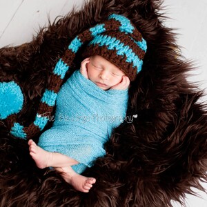 May include: A newborn infant wrapped in a light blue swaddle blanket, wearing a long, striped brown and light blue crochet hat with a matching light blue pom-pom at the end. The infant is resting on a dark brown faux fur textured material.