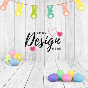 Könnte beinhalten: Ein weißer Holz Hintergrund mit einer Girlande aus pastellfarbenen Hasenschnitten, die von oben hängen. Der Text "Your Design Here" ist in schwarzer Kursivschrift geschrieben. Am unteren Rand des Bildes befinden sich mehrere bunte Ostereier.