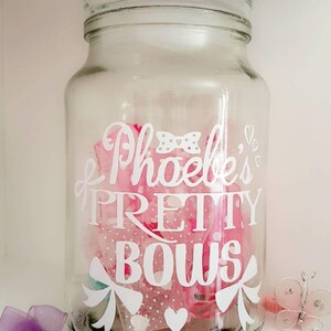 Może przedstawiać: Przezroczysty szklany słoik wypełniony różowymi kokardkami, z białym napisem "Phoebe's Pretty Bows". Słoik stoi na białej półce, z fioletową kokardką i dekoracyjnym motylem. Kokardki są w różnych odcieniach różu.