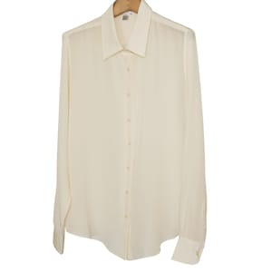 May include: Cream-colored long-sleeve button-down shirt hanging on a wooden hanger. The shirt has a classic collar, buttoned cuffs, and a tailored fit. The fabric appears to be lightweight and slightly sheer, suggesting a formal or professional style.