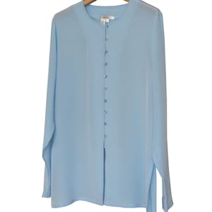 Könnte beinhalten: Hellblaue Langarmbluse mit Knopfleiste und rundem Ausschnitt. Die Bluse hängt an einem Holzkleiderbügel. Der Stoff wirkt leicht und fließend.