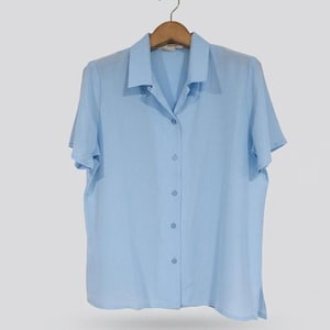 May include: A light blue short-sleeved button-down shirt with a classic collar. The shirt features a straight hemline with small side slits and a vertical row of matching light blue buttons running down the center front of the garment.