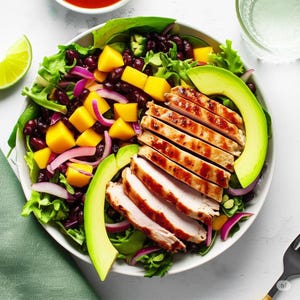 May include: A colourful salad in a white bowl, with grilled chicken, avocado slices, mango pieces, red onion, and black beans. The salad sits on a bed of green lettuce, with a lime wedge and a small bowl of red sauce visible.