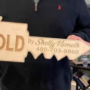 以下が含まれることがあります： 「SOLD」と「By Shelly Nemeth 480-703-8860」という茶色の文字が書かれた、木の鍵の形をした看板です。
