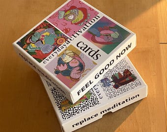 May include: Two white card boxes stacked on a wooden surface. The top box has the words "everyday activation cards" and "FEEL GOOD NOW" printed on it. The boxes have colorful abstract art.