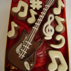 May include: A red box filled with chocolate musical notes and a chocolate guitar. The guitar is brown with white strings and a white pickguard. The notes are white with brown outlines.